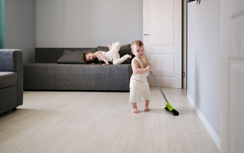  Toddler holding broom on light flooring, sibling resting on a couch.