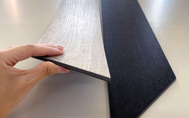 Loose lay flooring samples showing the front with a light wood grain and the back with a non-slip black layer.