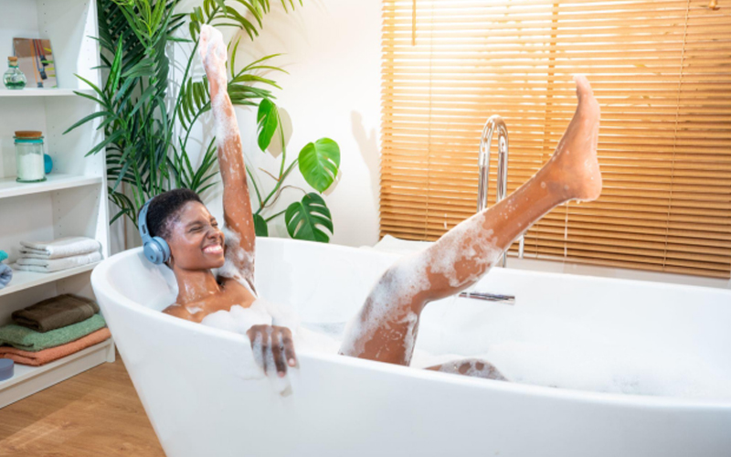 Elegant Black woman enjoying music with headphones in a foam-filled bathtub, set on wood-look vinyl flooring.